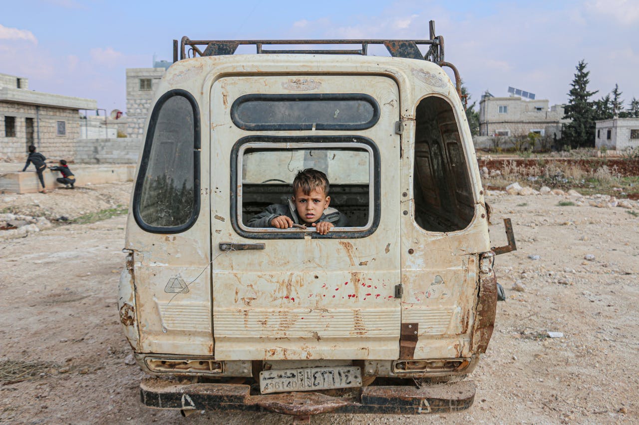 services-04 A young boy peers from an abandoned van in the desolate streets of Idlib, Syria, amidst war devastation.